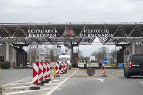 Republic of Bulgaria official border crossing entry gate with national coat of arms, Bulgarian and EU flags, border officer inspecting vehicles with red and white barriers
